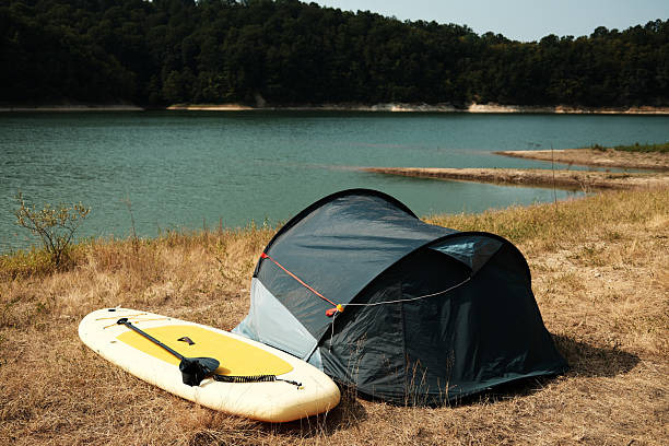 10 Model Tenda Camping Aesthetic yang Paling Instagrammable papan sup kuning tiup dan tenda berkemah biru di atas rumput kering dekat danau dengan latar